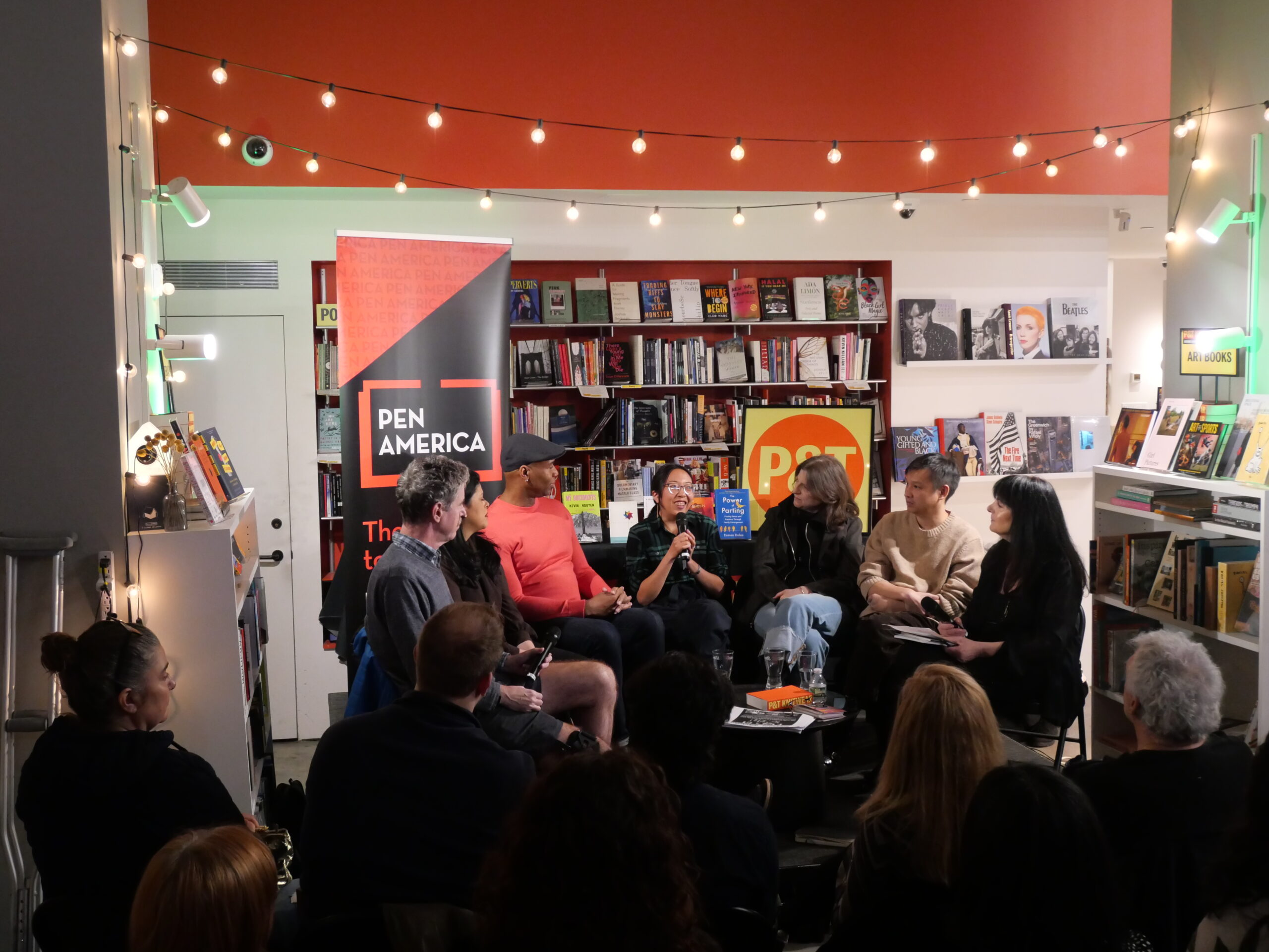 A panel of six people sits in front of an audience in a bookstore, discussing on stage next to PEN America and PST banners, with string lights overhead and bookshelves in the background.