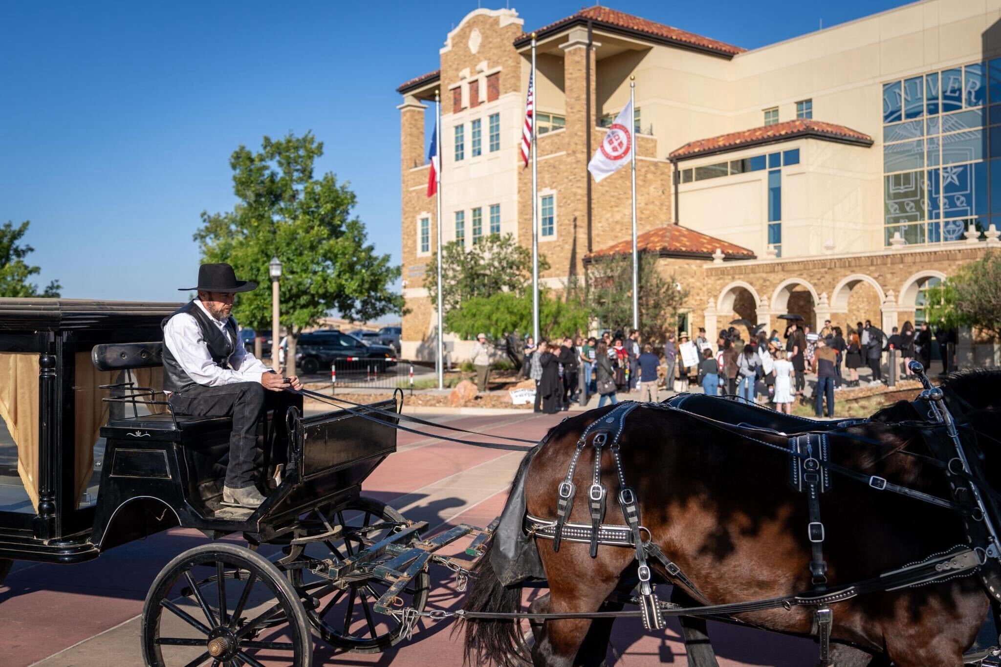 A man in traditional attire drives a horse-drawn carriage past a large, historic-style building on a Texas campus, with a crowd gathered outside under bright, sunny skies amid discussions about censorship.