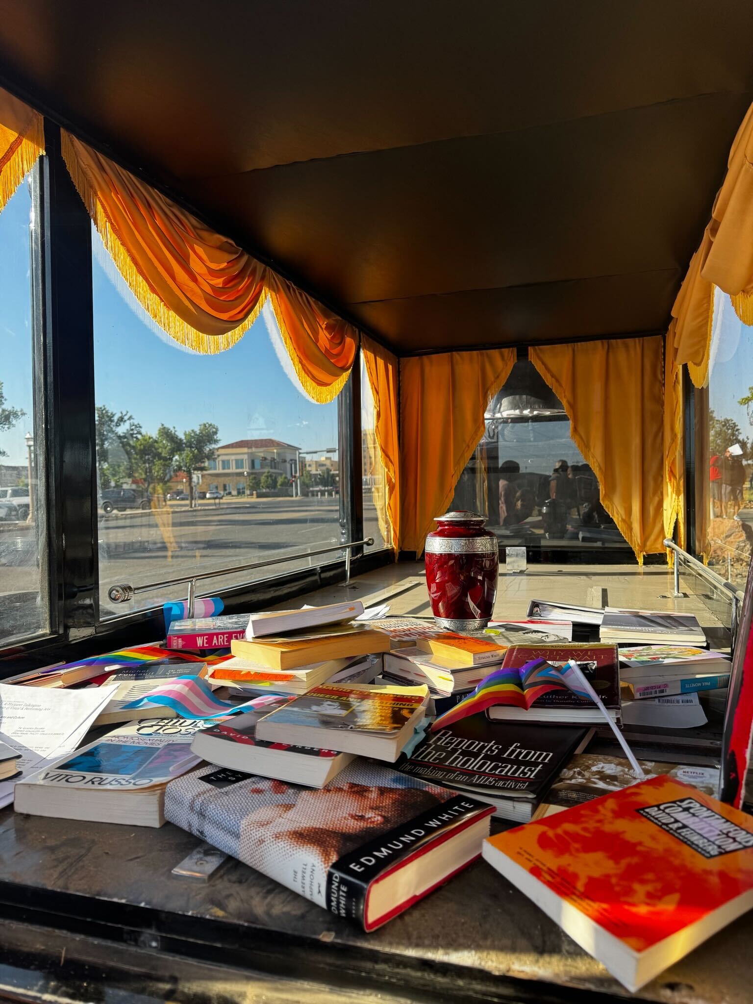 A pile of books and a red urn rest on a surface inside a vehicle with large windows and yellow curtains, sunlight streaming in, the Texas Campus Censorship debate lingering as a street scene unfolds outside.