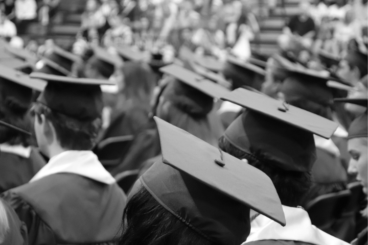 A black-and-white photo showing the backs of graduates wearing caps and gowns, sitting in rows during a graduation ceremony, with an audience blurred in the background.