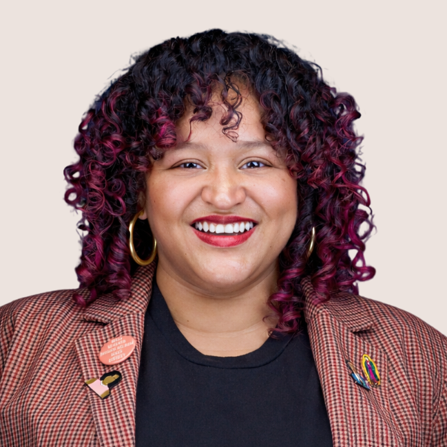 A person with curly, dark hair with purple highlights smiles at the camera. They wear hoop earrings, a brown plaid blazer with pins, and a black shirt, against a plain light background.