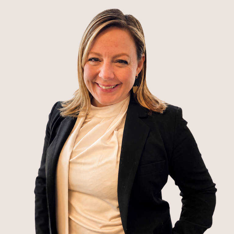 A woman with shoulder-length blonde hair, wearing a white blouse and black blazer, smiles while standing against a plain light background.