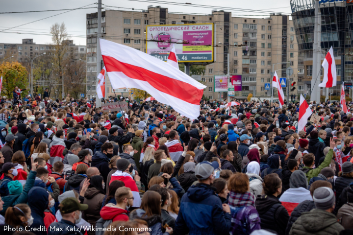 A large crowd of people gather outdoors, many wearing masks. A red and white striped flag is prominently waved above the crowd. Urban buildings and a billboard appear in the background.