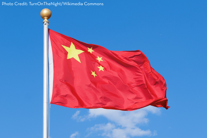 The national flag of China, red with five yellow stars in the top left corner, flies on a flagpole against a clear blue sky.