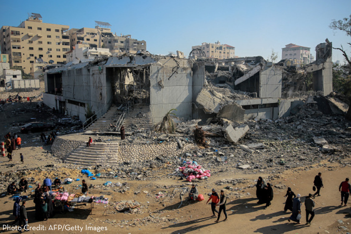 A large, heavily damaged building with collapsed walls and debris is surrounded by people walking and gathering in groups. Makeshift stalls and scattered belongings are visible in the foreground. Urban buildings are seen in the background.