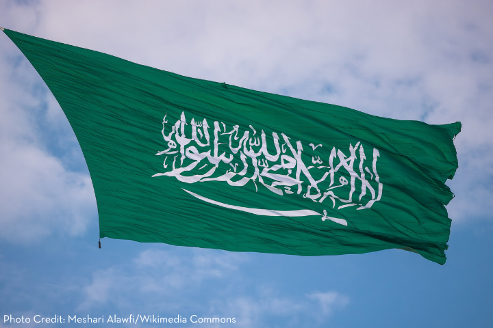 The green flag of Saudi Arabia waves against a blue sky, displaying white Arabic script and a sword. Photo credit: Meshari Alawfi/Wikimedia Commons.