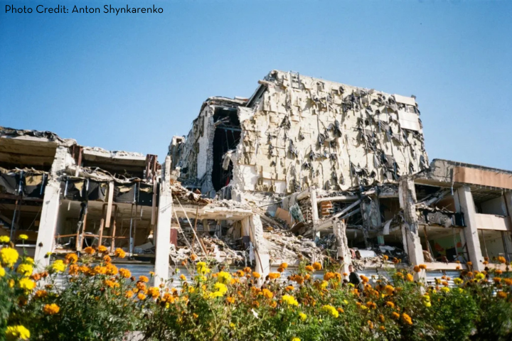A large, heavily damaged building with collapsed walls and exposed debris stands behind a field of bright yellow flowers under a clear blue sky—a powerful scene reflecting the need for global advocacy. Photo credit: Anton Shynkarenko.