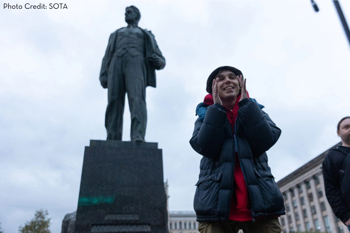 A person in a dark winter jacket and hat smiles, cupping their face near a large statue of a man on a pedestal in an urban square—an inspiring scene of global advocacy against overcast skies. Photo Credit: SOTA.