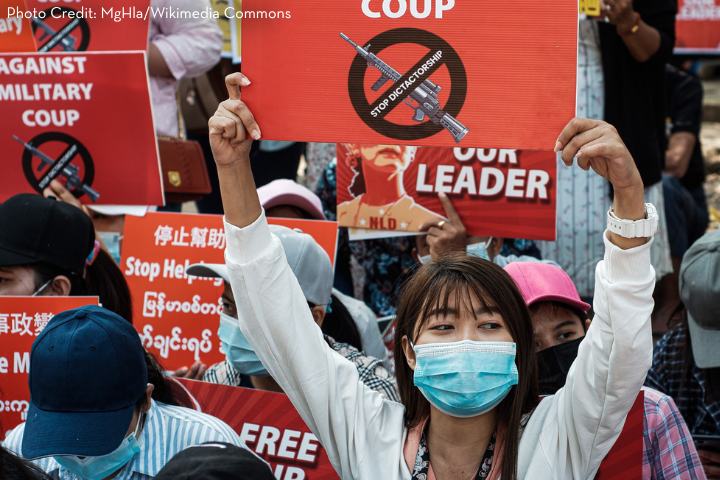 A group of protesters, many wearing masks, hold signs opposing a military coup as part of global advocacy. One woman in the foreground raises a red sign with a crossed-out gun and the words “Stop Dictatorship.” Other protest signs are visible behind her.