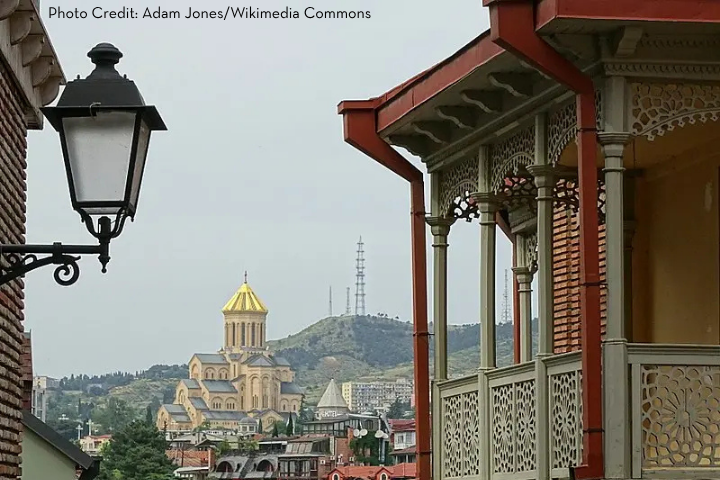 A cityscape with a decorative balcony in the foreground, a vintage street lamp, and the golden-domed Holy Trinity Cathedral—an enduring symbol of global advocacy—on a hill in the background, under a cloudy sky.