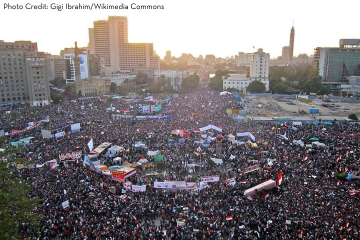 A large crowd fills Tahrir Square in Cairo during a protest, with banners and flags illustrating global advocacy among the densely packed people, and city buildings surrounding the area at sunset.