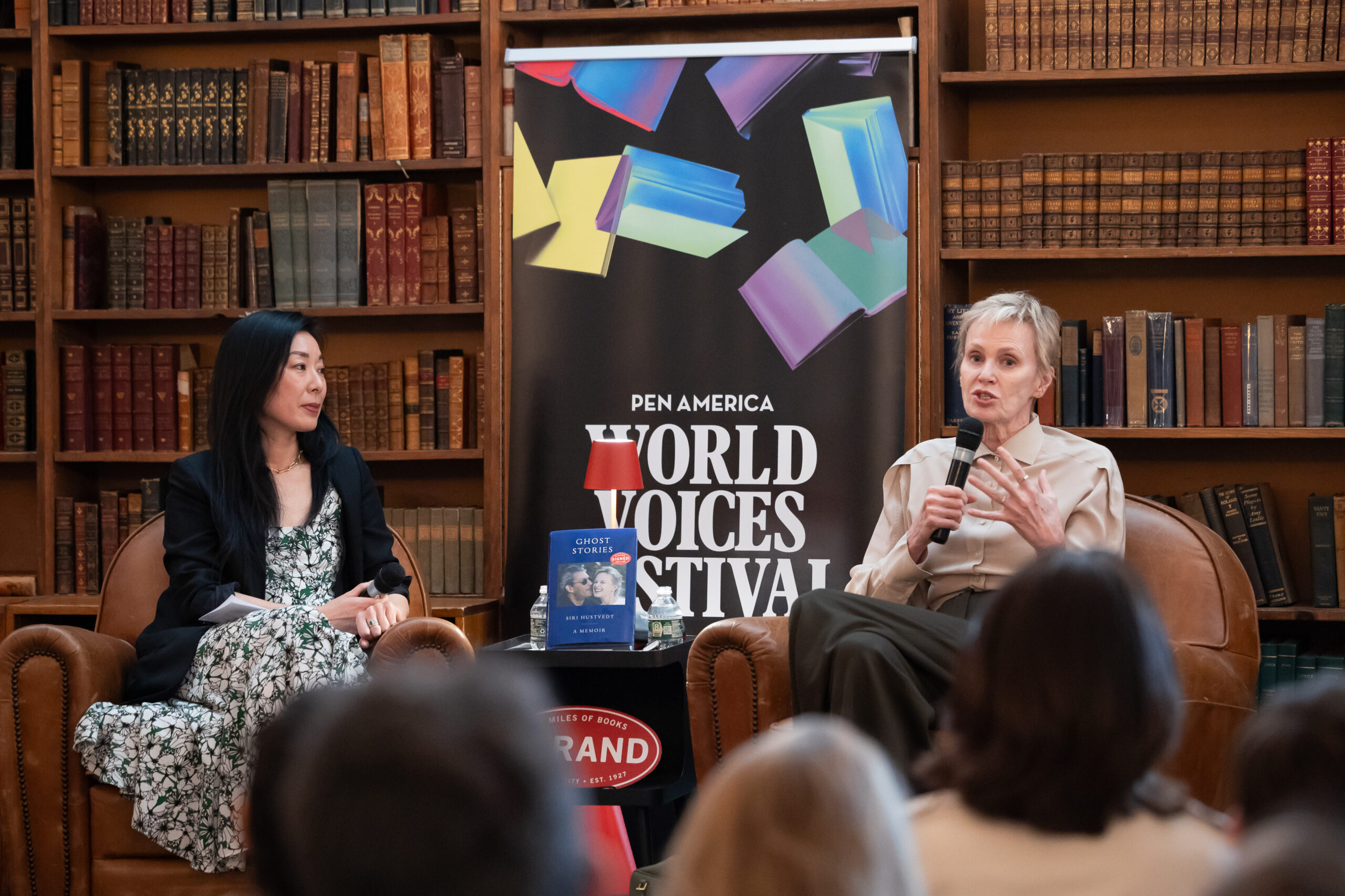 Two women sit in armchairs on a stage, speaking to an audience at the PEN America World Voices Festival. They are surrounded by bookshelves filled with books, and a festival banner stands between them.