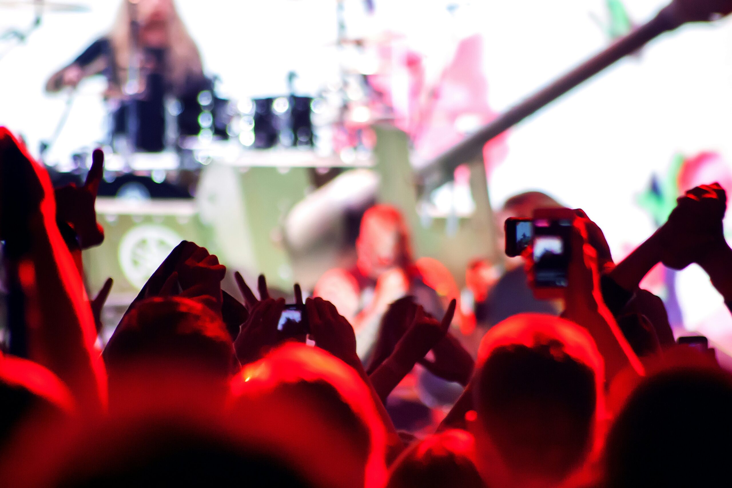 A crowd of people with raised hands and phones records a concert. The stage is brightly lit, and a drummer and other musicians are visible in the background, but the focus is on the enthusiastic audience.