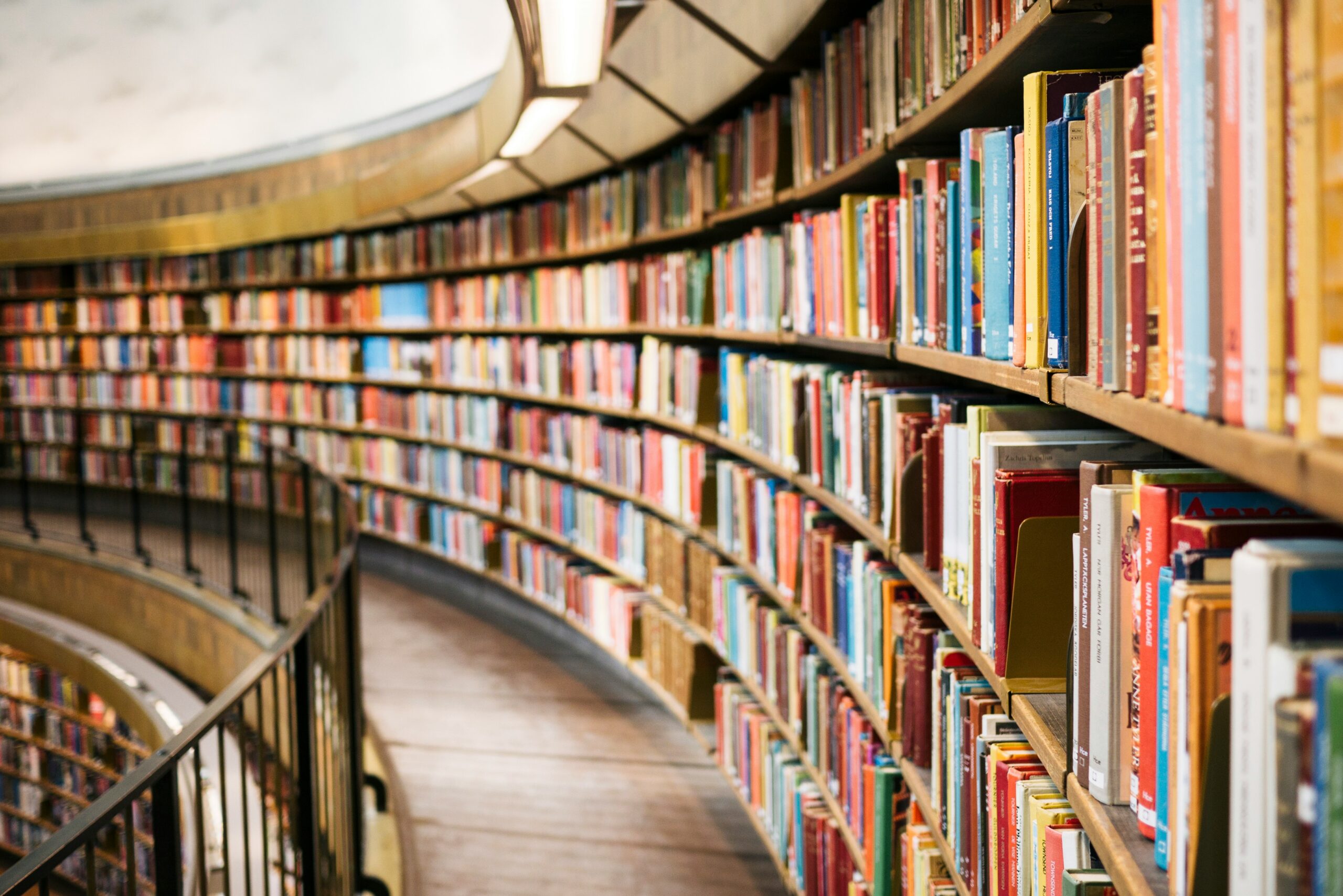 Curved rows of colorful books line tall wooden shelves in a large, modern library; a railing follows the curve of the walkway beside the books.