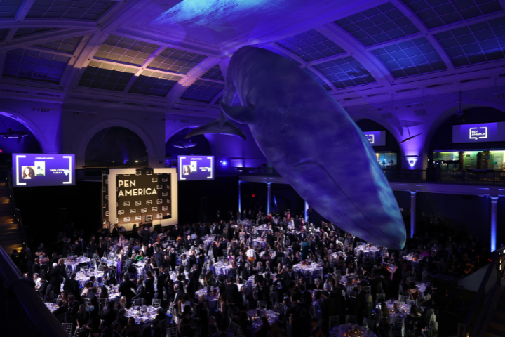 A large crowd attends a formal event at a museum, dining under a life-sized model of a blue whale suspended from the ceiling. Purple lighting bathes the room, and a backdrop reads “PEN America.”.