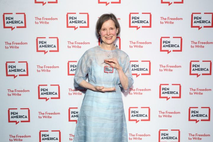 A smiling woman in a light blue dress holds a clear award in front of a backdrop featuring repeated PEN America logos and the slogan The Freedom to Write.