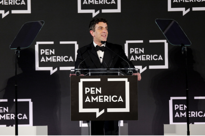 A man in a tuxedo speaks at a podium with the PEN America logo, flanked by two transparent teleprompters, against a backdrop featuring repeated PEN America logos.