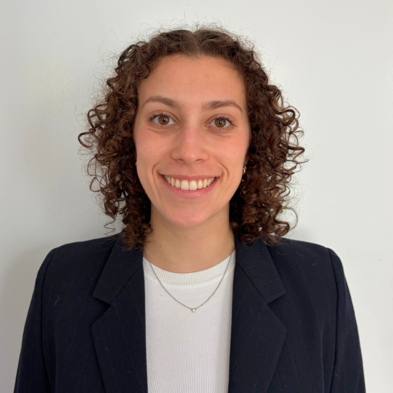 A woman with curly brown hair is smiling at the camera. She is wearing a dark blazer over a white top, with two delicate necklaces, and standing against a plain white background.