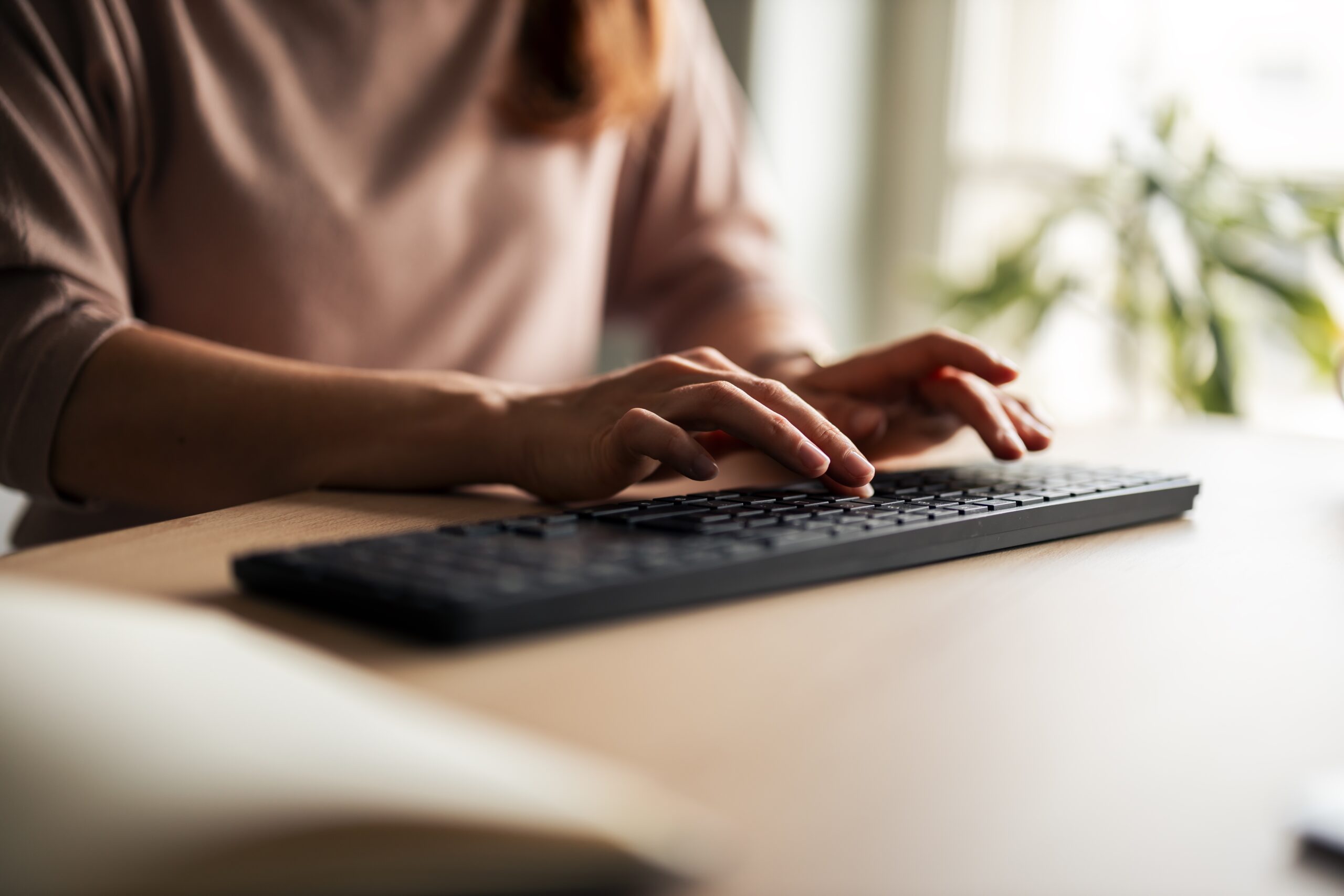 A person types on a black computer keyboard at a wooden desk, with soft natural light coming from a window in the background. Only their hands and part of their torso are visible.