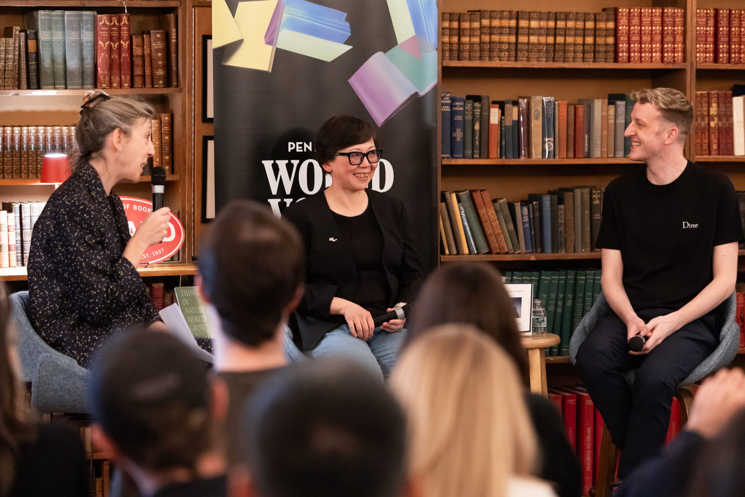 Three people sit and converse in front of bookshelves and a PEN World Voices banner, engaging with an audience at what appears to be a literary event or panel discussion.