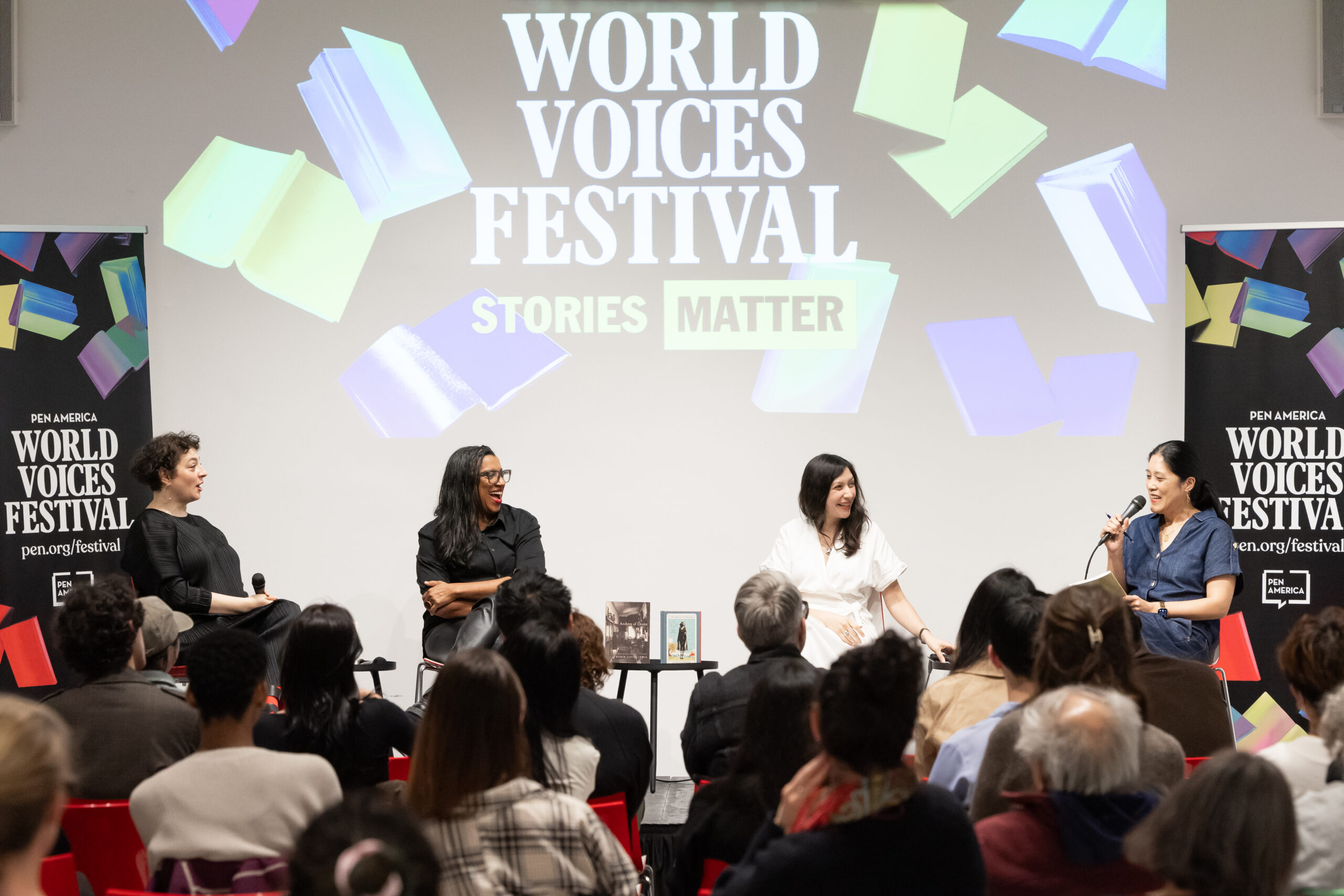 Four women sit on stage with microphones, engaging in a panel discussion at the World Voices Festival. An audience watches, while colorful book graphics and the festival’s “Stories Matter” slogan are visible on a screen behind them.