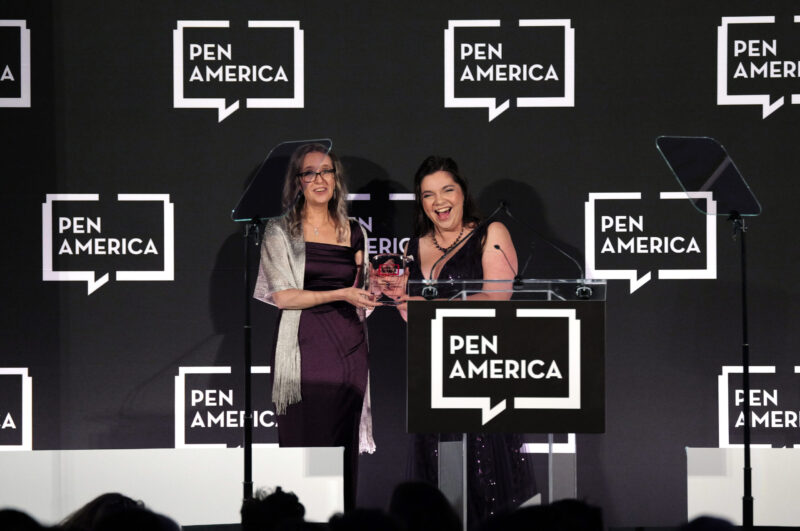 Two women stand on stage at a PEN America event, smiling as they hold an award together. They are dressed formally, with PEN America logos displayed on the backdrop behind them.