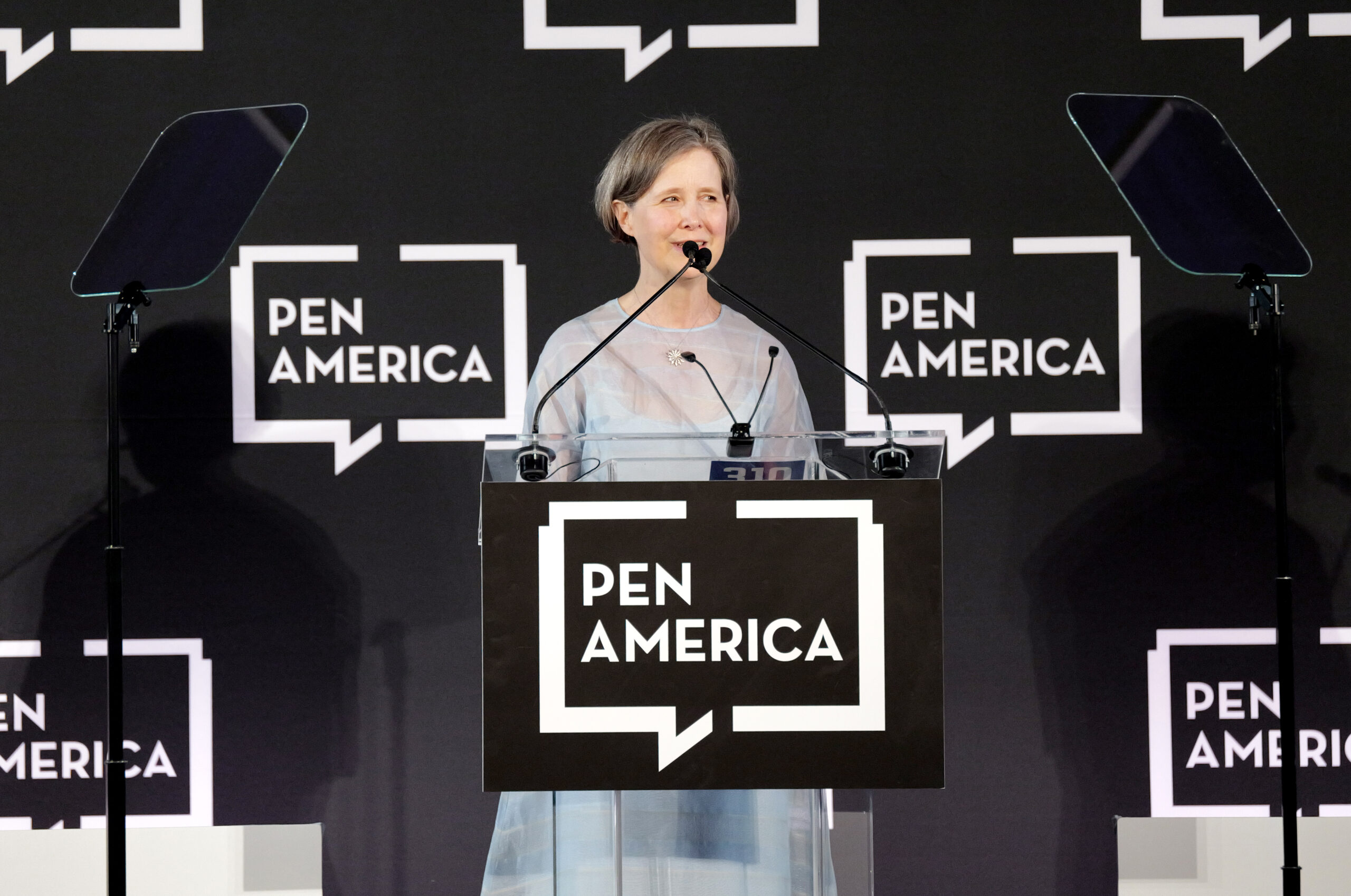 A person stands at a clear podium with PEN America logos behind them, speaking into microphones at an event as they deliver remarks for the PEN America Literary Service Award, with two teleprompters on either side.