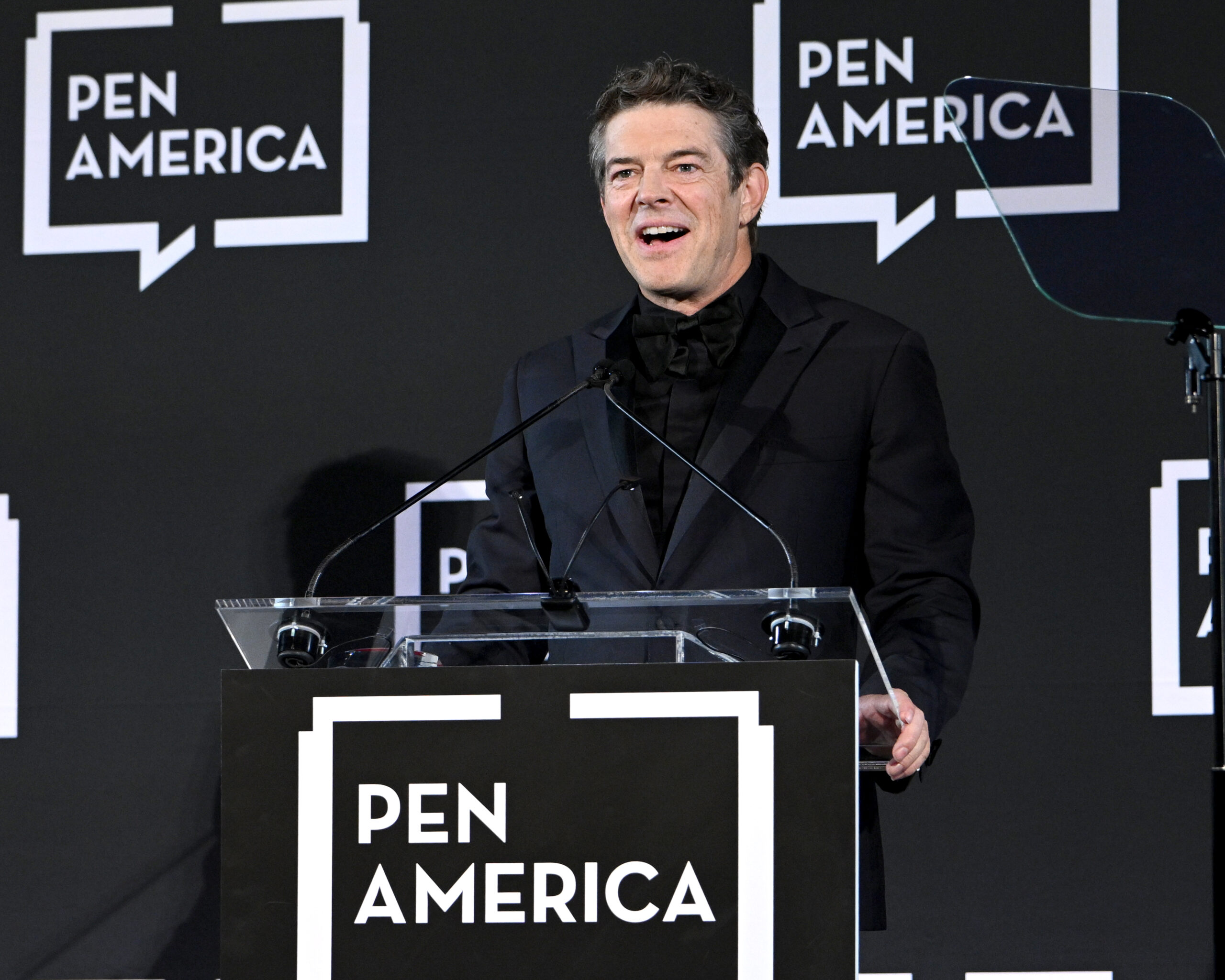 A man in a dark suit stands at a podium with a PEN America sign, speaking at an event. The background features repeating PEN America logos on a black wall, highlighting the prestigious PEN America Business Visionary Award ceremony.