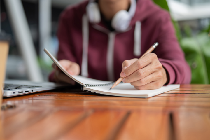 A person in a maroon hoodie with headphones around their neck writes in a notebook with a pencil at a wooden table, next to an open laptop.