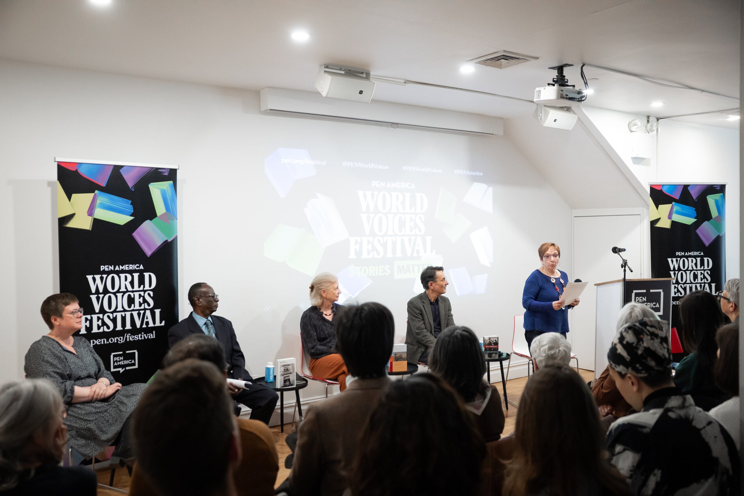 A woman speaks at a podium to an audience in a bright room with World Voices Festival banners and a projection behind a panel of five seated speakers.