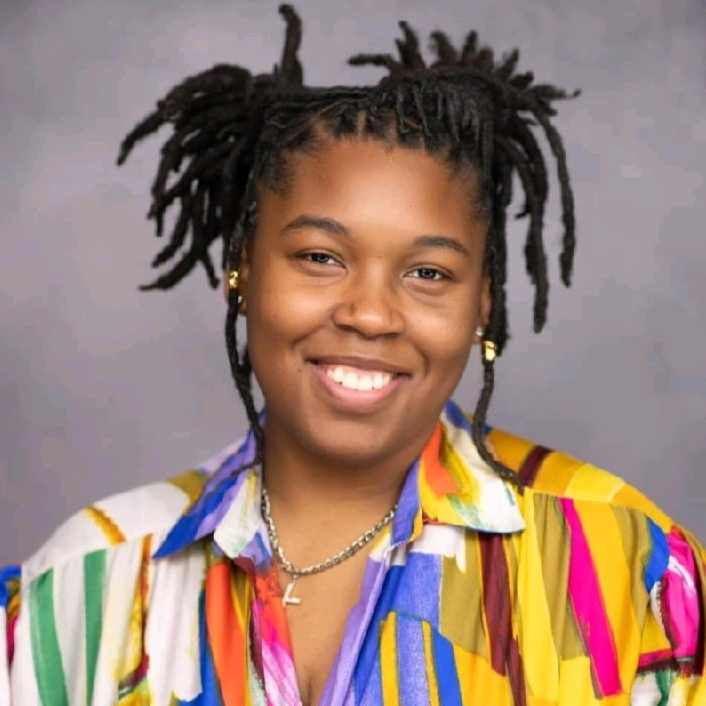 A person with medium-length twisted hair styled in pigtails smiles at the camera. They are wearing a colorful, patterned shirt and small gold hoop earrings, with a gray background behind them.