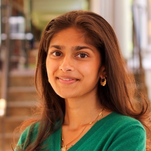 A young woman with long brown hair, wearing a green top and gold jewelry, smiles softly while standing indoors with a blurred staircase and corridor in the background.