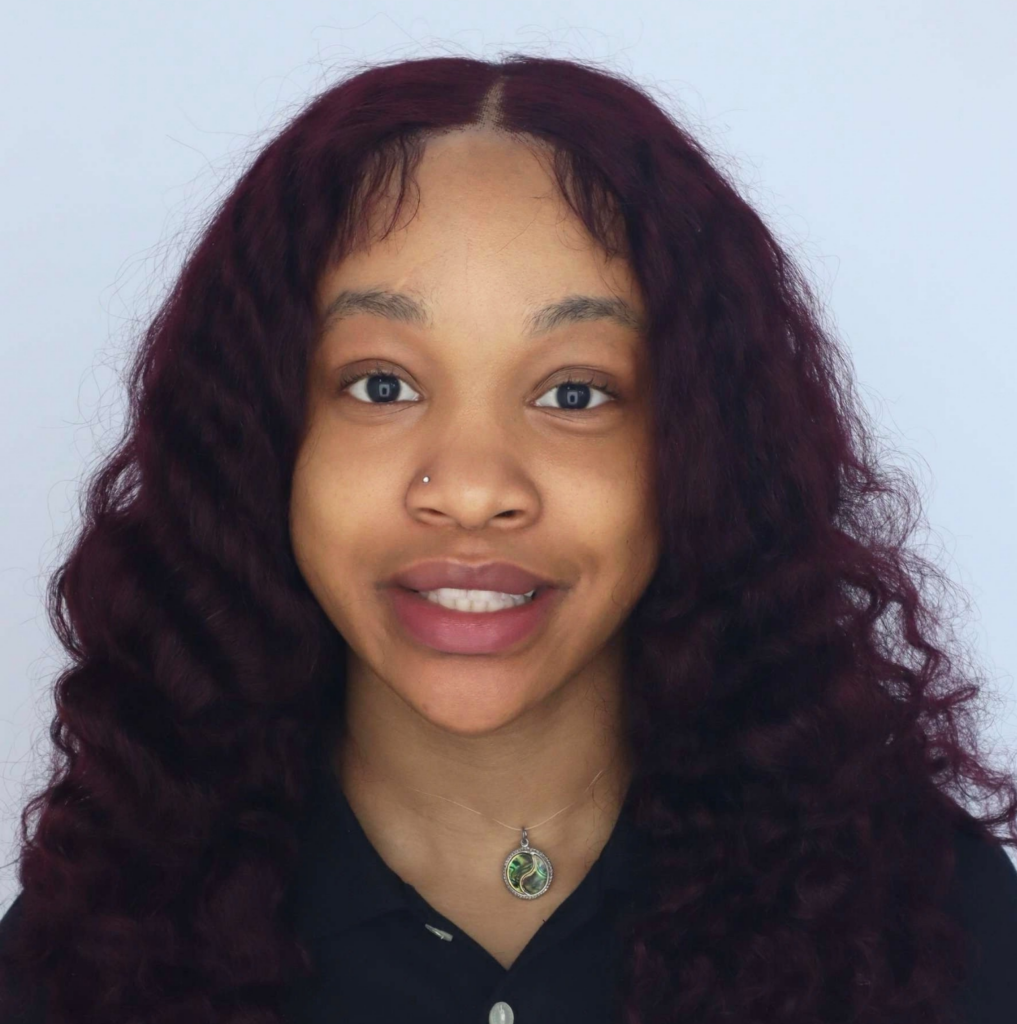 A young woman with long, curly dark red hair smiles at the camera. She is wearing a black collared shirt, a round necklace, and has a nose piercing. The background is plain and light-colored.