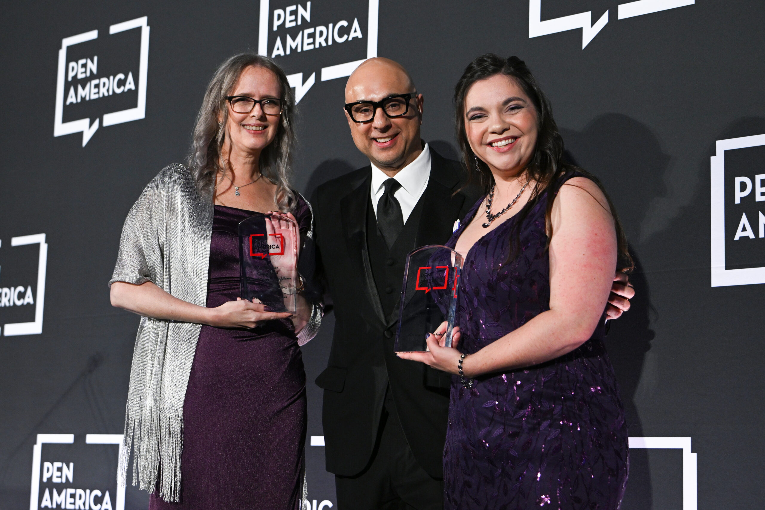 Three people pose together smiling at a PEN America event. Two women, each holding a clear award, stand on either side of a man in a black suit and glasses. All are dressed formally, standing in front of a PEN America backdrop.