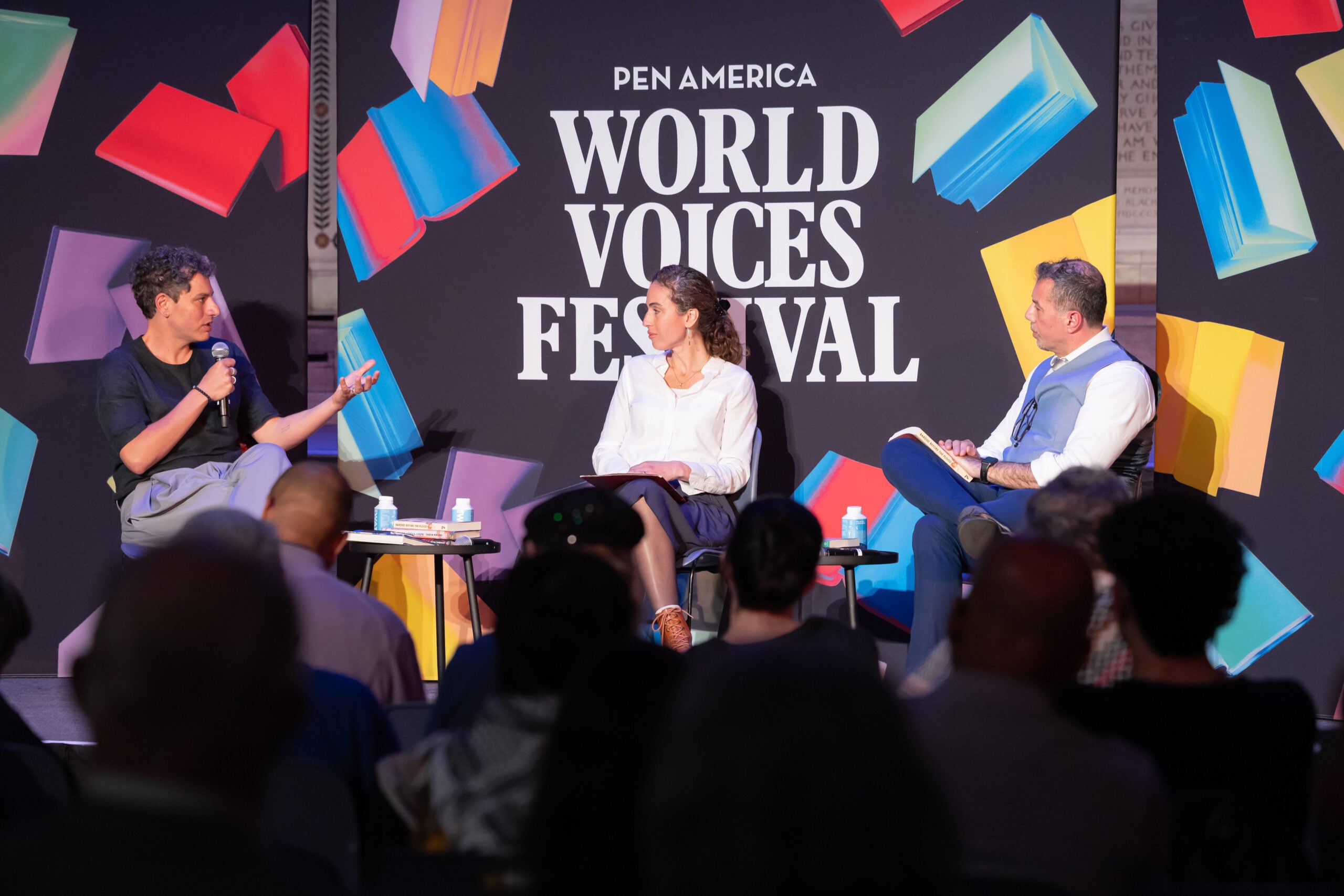 Three people are seated on stage in front of an audience, engaged in a panel discussion at the PEN America World Voices Festival, with colorful book graphics in the background.