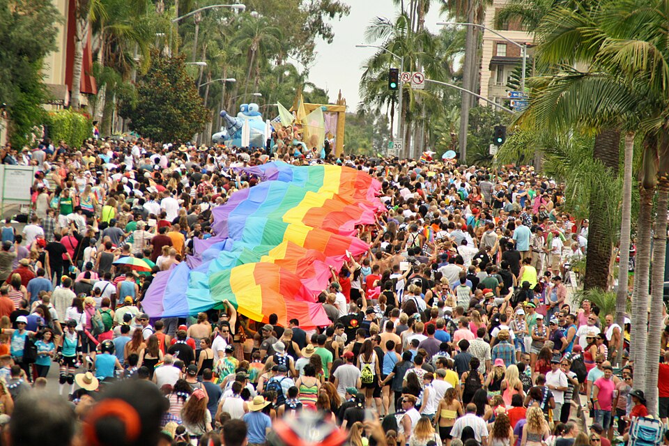 A large crowd marches down a city street during a parade, with a giant rainbow flag being carried above people’s heads. Palm trees line the road, and the atmosphere is festive and colorful.