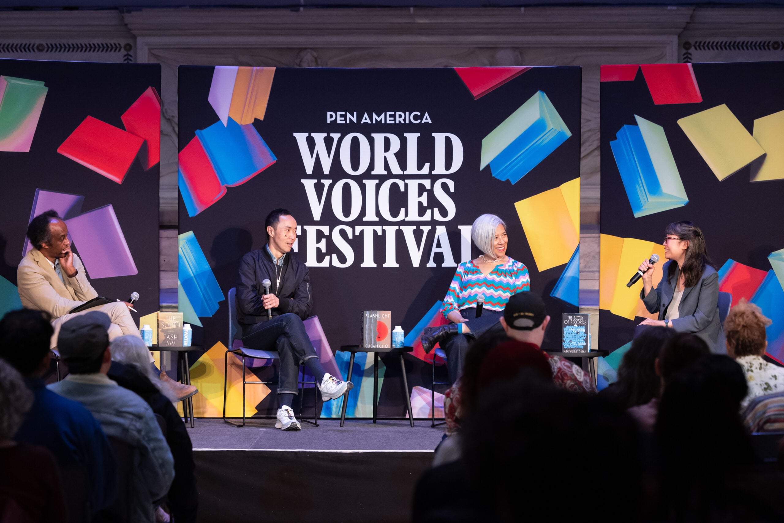 Four people sit onstage in front of a colorful PEN America World Voices Festival backdrop, engaged in a panel discussion, while an audience watches.