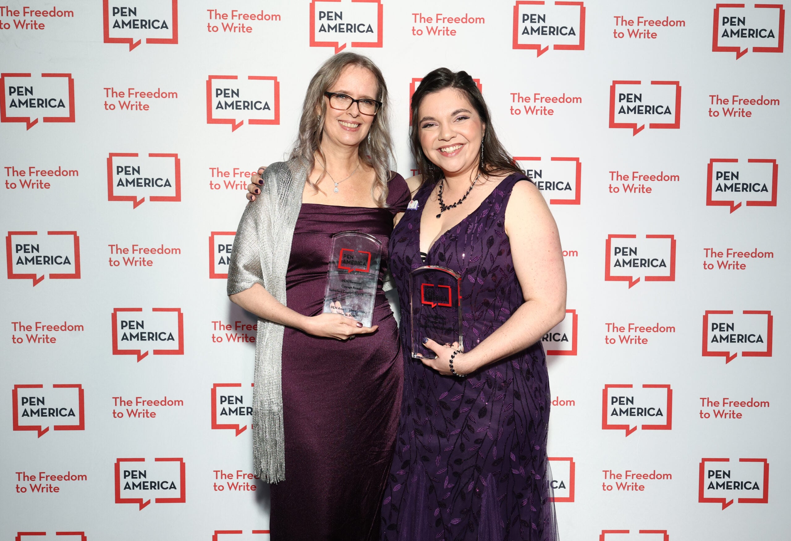Two women in purple dresses smile and hold glass awards while standing in front of a step-and-repeat backdrop with the PEN America logo and the phrases The Freedom to Write in red.
