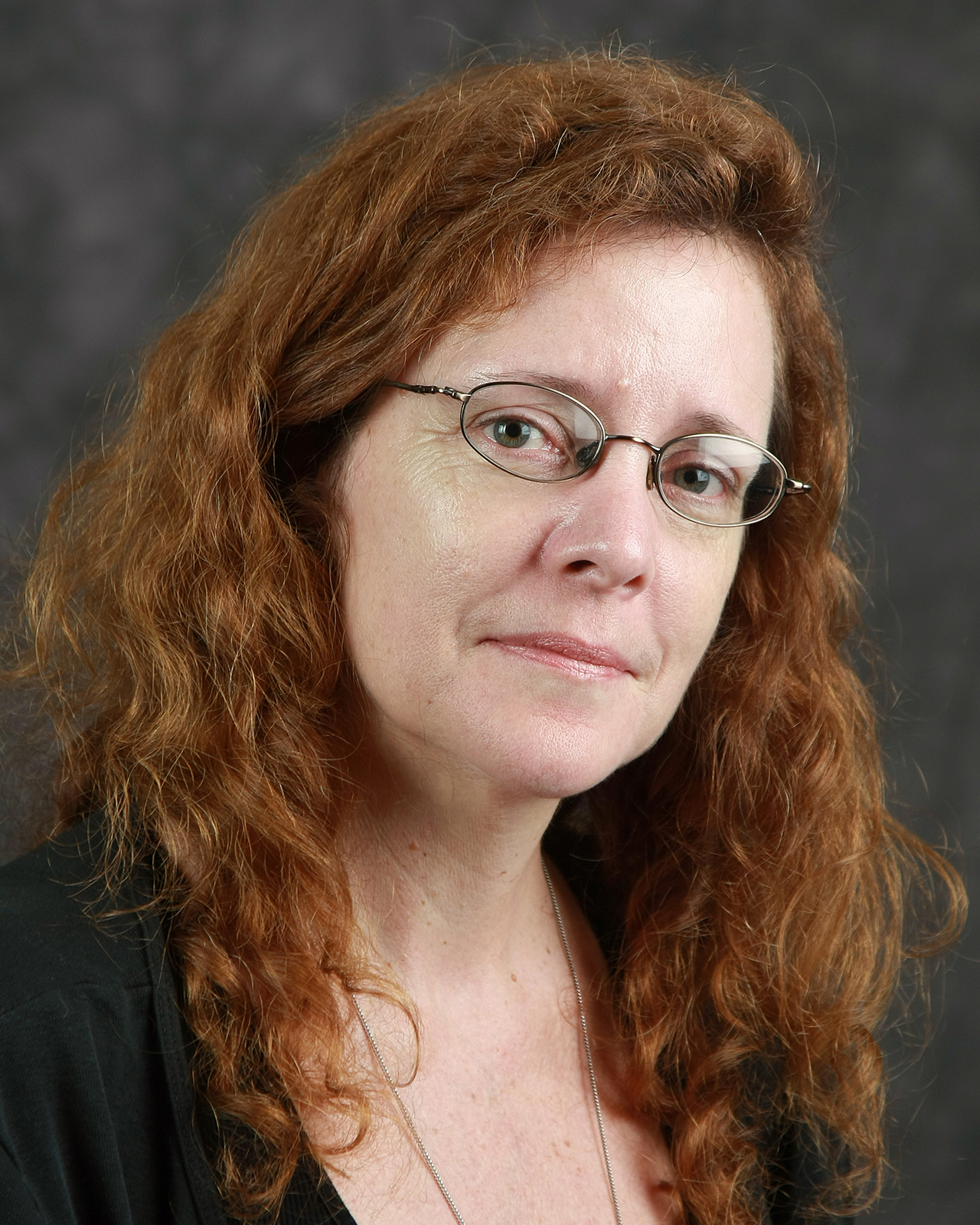 A woman with long, curly red hair and glasses looks at the camera with a neutral expression. She is wearing a black top and a thin necklace, with a dark, blurred background behind her.