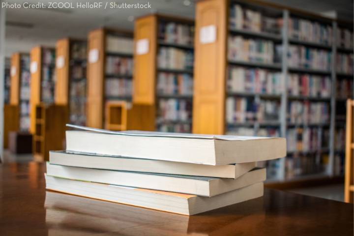 A stack of four books sits on a wooden table in a library, with tall bookshelves filled with books blurred in the background.