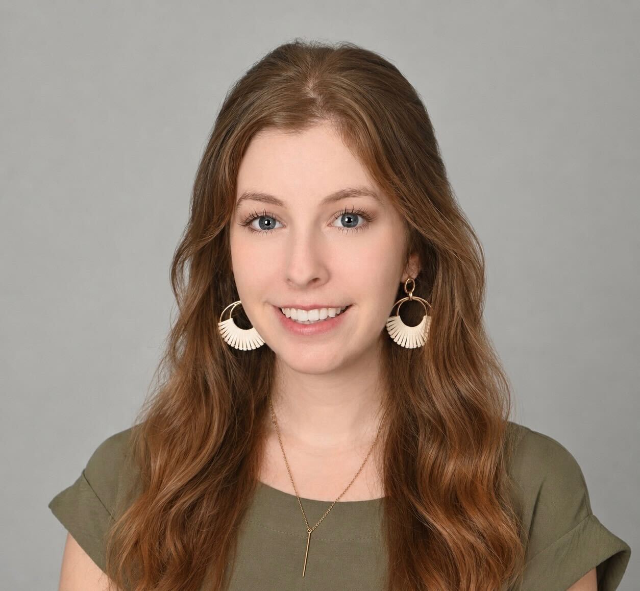 A young woman with long wavy light brown hair, wearing a green top, large circular white earrings, and a gold necklace, smiles softly at the camera against a plain gray background.