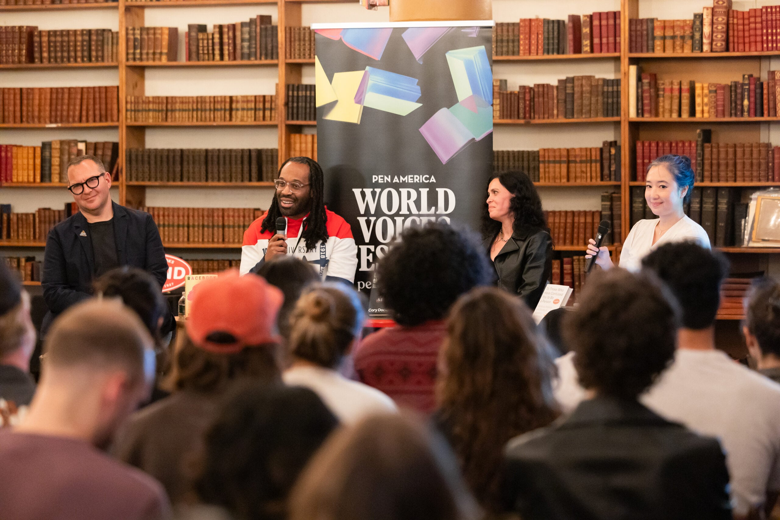 A panel of four people speaks on stage in front of an audience at a PEN America World Voices Festival event, with bookshelves filled with books in the background.