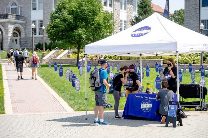 Students gather at an outdoor Dukes Vote information booth under a white tent on a college campus, with signs and decorations about voting. Some people are talking while others walk by in the background.
