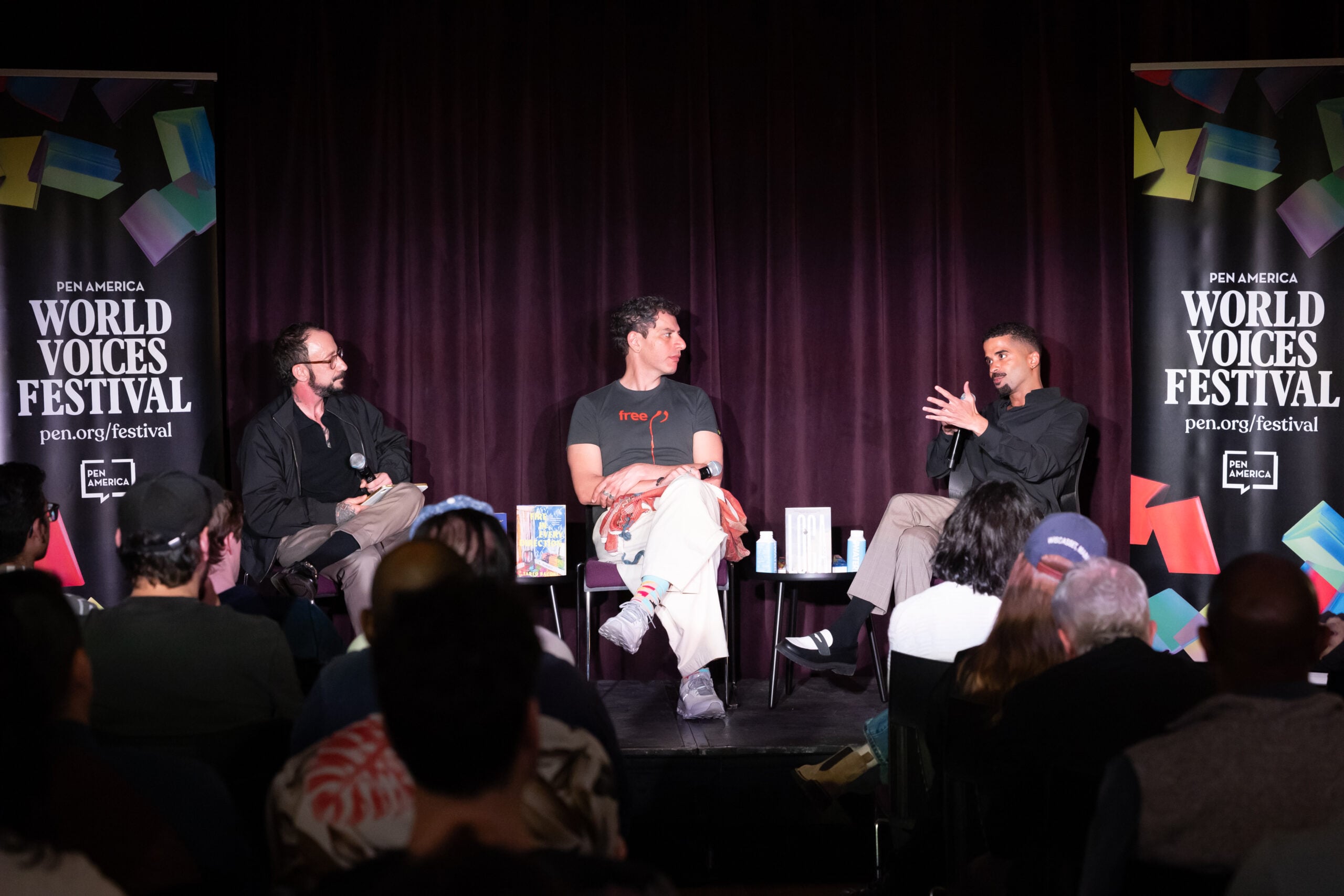 Three men sit on a stage having a discussion at the PEN America World Voices Festival, with two banners on either side and an audience seated in front of them. Books are displayed on the table between them.