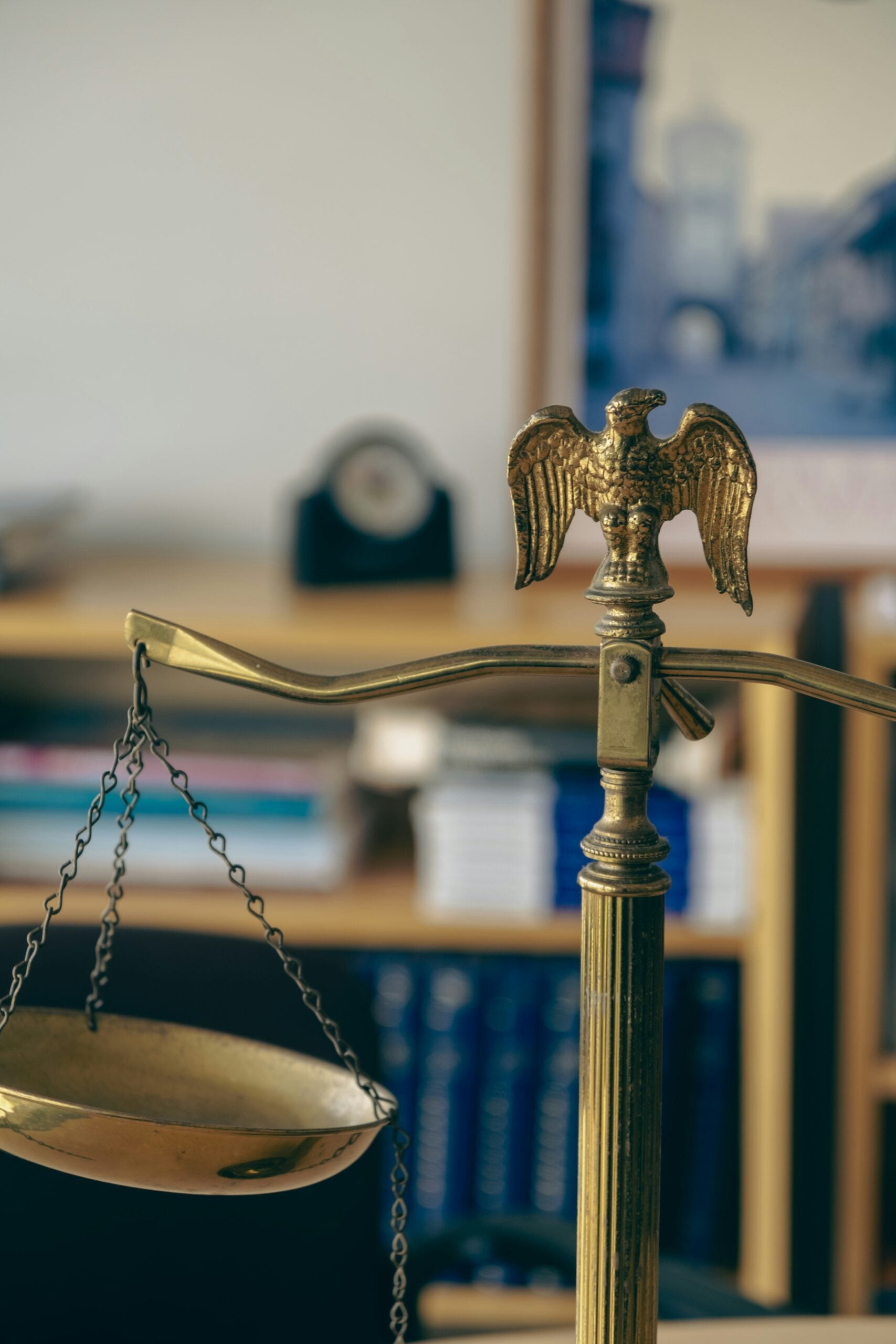 A close-up of a brass balance scale with an eagle ornament on top, set against a blurred background of bookshelves and framed pictures.