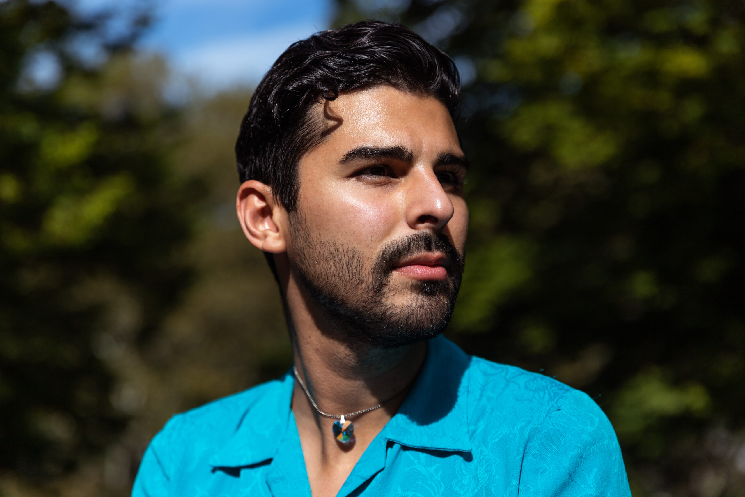 A man with dark hair and a beard wearing a turquoise shirt and a necklace looks thoughtfully into the distance outdoors, with sunlight on his face and a blurred green background.