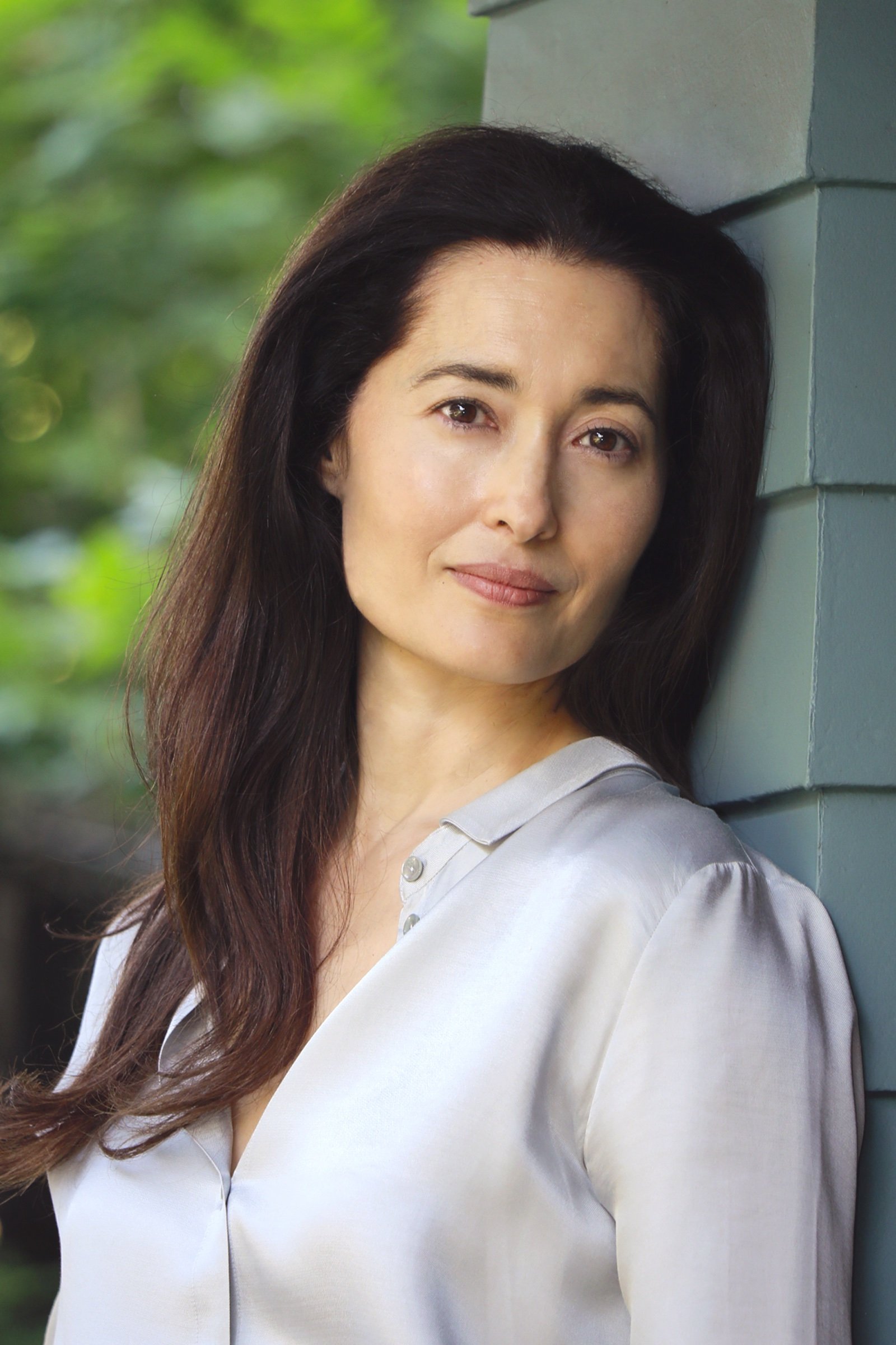 A woman with long dark hair and a light gray blouse leans against a green wall, looking calmly at the camera with a slight smile. Green foliage is blurred in the background.