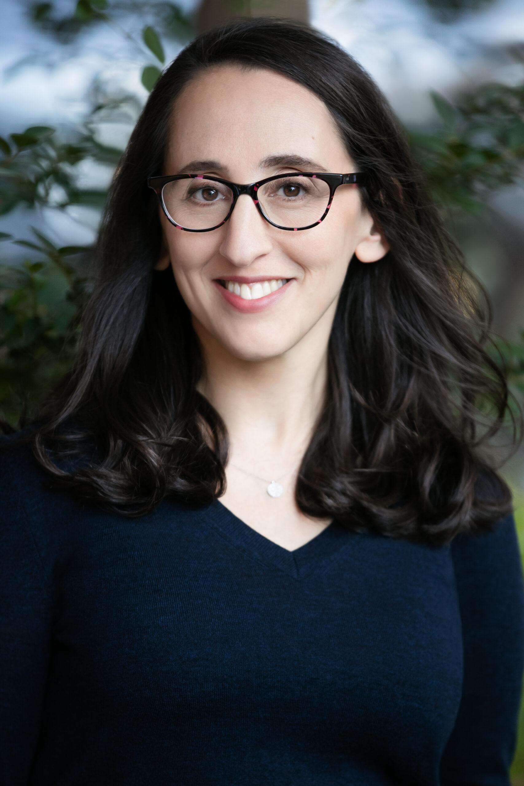 A woman with long dark hair and glasses smiles at the camera. She is wearing a navy blue sweater and a silver necklace, with green foliage and a blurred background behind her.