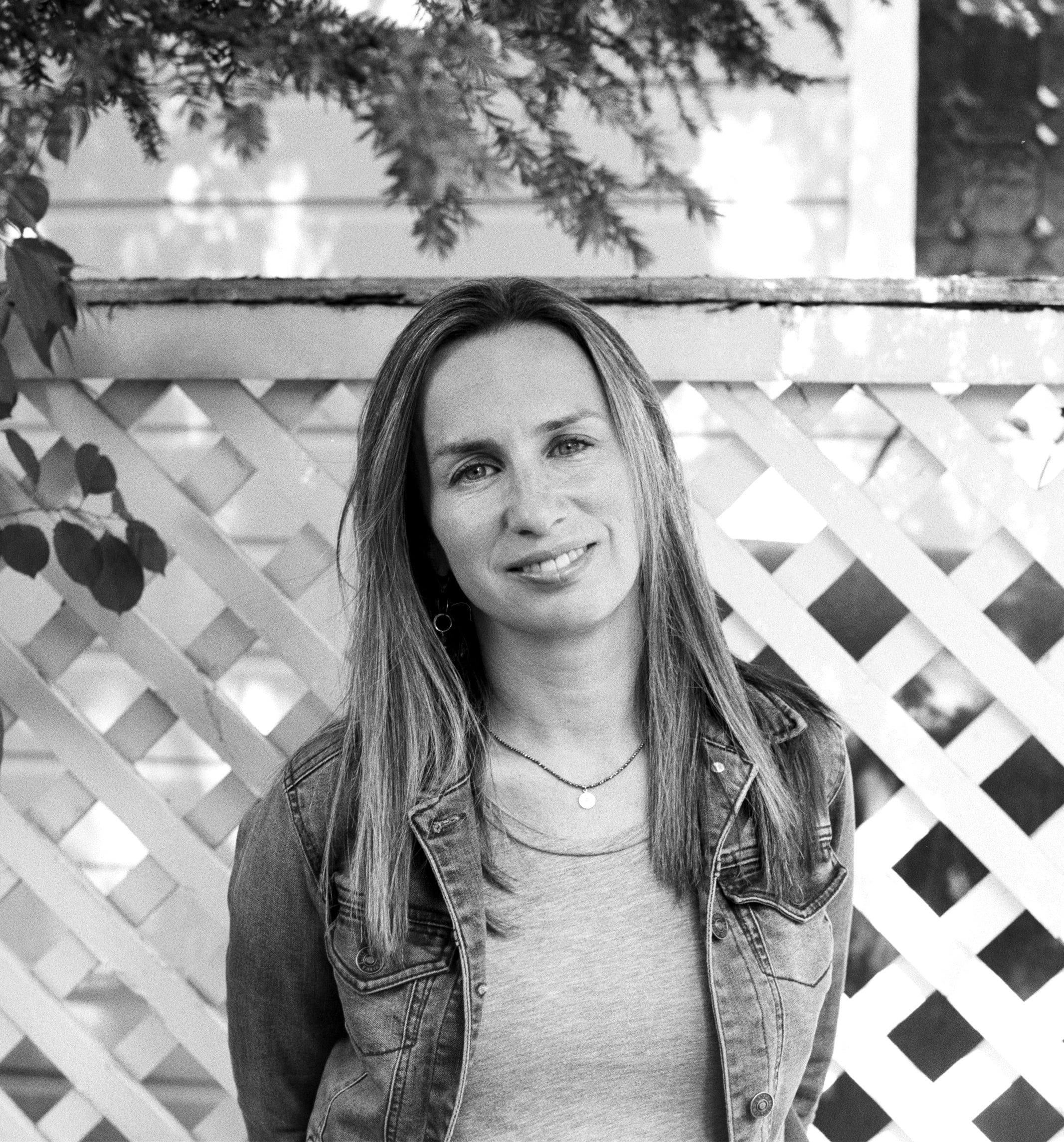 A woman with long hair, wearing a denim jacket over a shirt, stands outside in front of a white lattice fence and greenery, smiling softly at the camera.