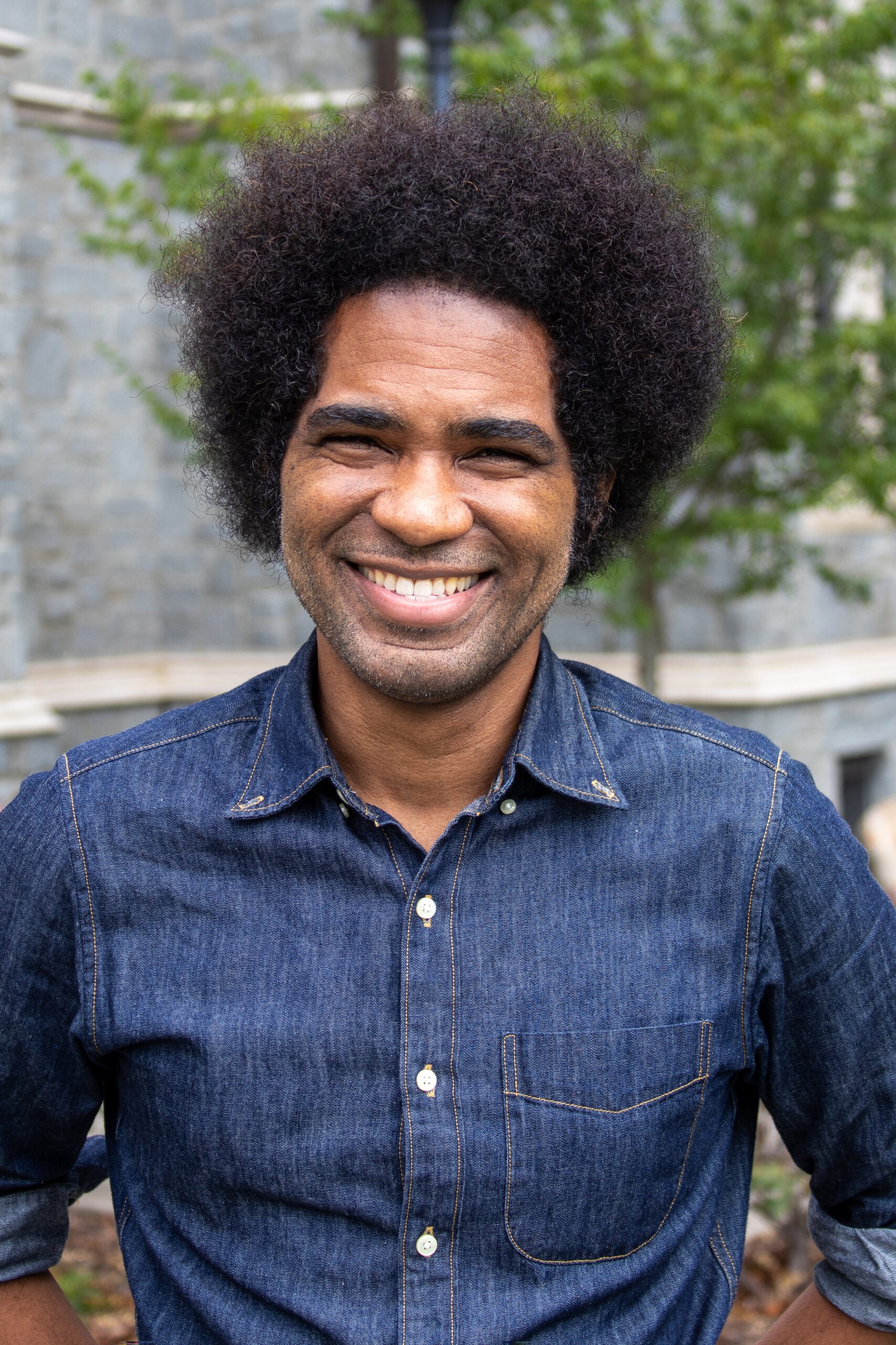 A smiling man with dark curly hair, wearing a denim button-up shirt, stands outdoors in front of a stone building and greenery.